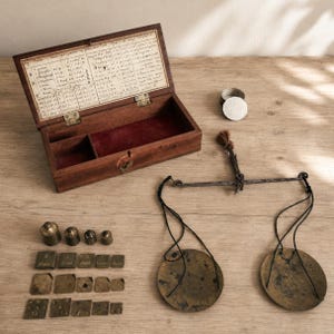 Peut inclure: Une boîte en bois ancienne avec un intérieur en velours rouge et un tableau de conversion imprimé sur le couvercle, accompagnée d'une balance en métal à deux plateaux circulaires et d'une collection de poids en laiton et métal variés.