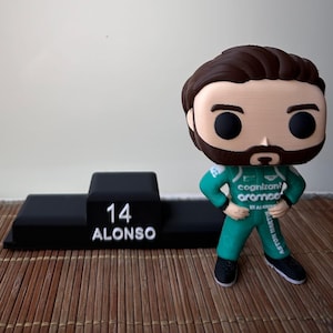 Puede incluir: Figura Funko Pop de un piloto de Fórmula 1 con un traje de carreras verde, junto a un podio negro con el número 14 y el nombre ALONSO. La figura tiene barba y pelo castaño.