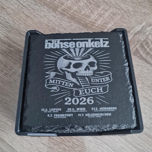 Könnte beinhalten: Ein schwarzes Schiefer-Untersetzer-Set mit dem Text "böhse onkelz" und einer Totenkopf-Grafik. Der Untersetzer enthält auch den Text "MITTEN UNTER EUCH 2026" sowie mehrere Städtenamen und Daten.