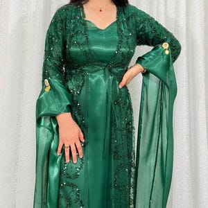 Könnte beinhalten: Ein bodenlanges smaragdgrünes Kleid mit einem satinartigen Oberteil und einem passenden transparenten Überwurf. Der Überwurf ist mit aufwendigen dunkelgrünen Paillettenstickereien und langen, fließenden Ärmeln verziert. Goldfarbene runde Verzierungen akzentuieren die Ärmelbündchen.