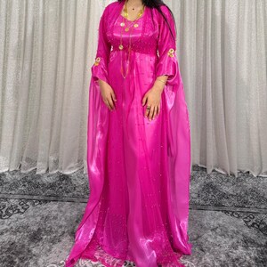 Könnte beinhalten: Ein bodenlanges, fuchsiafarbenes Kaftankleid mit langen, fließenden Ärmeln und einem transparenten Überwurf. Das Kleidungsstück ist mit zarten goldfarbenen Perlen und einer passenden goldfarbenen Halskette verziert, die über der Brust liegt.