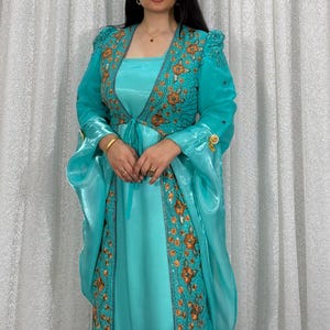 Könnte beinhalten: Ein helltürkisfarbenes, bodenlanges Kleid mit einem passenden, transparenten Überwurf, der vorne offen ist und lange, weite Ärmel hat. Der Überwurf ist mit goldener Blumenstickerei und kleinen Pailletten verziert und hat eine Bindung.