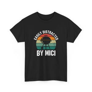 May include: Black t-shirt with a retro design featuring a grill illustration inside a striped circle. The text reads "EASILY DISTRACTED" above and "BY MICI" below the grill graphic. A casual tee for grilling enthusiasts.
