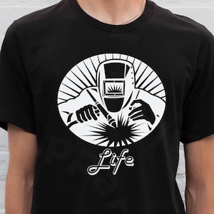 May include: Black t-shirt with a white graphic of a welder and the word "Life". The design is within a white oval with sunburst rays. The shirt is a crew neck. The person wearing the shirt is visible from the chest down.