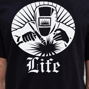 May include: Black t-shirt with a white graphic of a welder within a circular design. The graphic shows a welder wearing a mask and holding a welding torch. The word "Life" is written in a decorative font beneath the image.