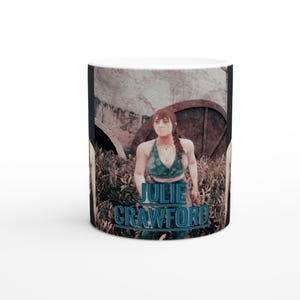 May include: White ceramic mug featuring a woman in a teal top and the text "JULIE CRAWFORD" in blue. The background includes a blurred image of a building and foliage, creating a unique design.