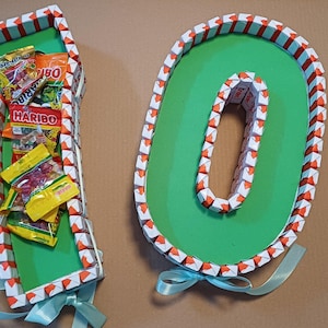 Puede incluir: Un número decorativo 10 hecho de cartón, con un interior verde y un borde de pequeños cuadrados coloridos. El número 1 está lleno de paquetes de caramelos. Una cinta azul claro está atada en la parte inferior.