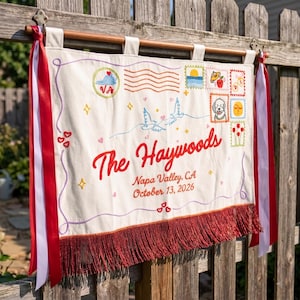 Könnte beinhalten: Besticktes Banner mit dem Text "The Haywoods" in roter Schrift, zusammen mit Datum und Ort. Das Banner zeigt verschiedene gestickte Designs, darunter Briefmarken, Tauben und Herzen, und ist mit roter Fransen und Bändern verziert.