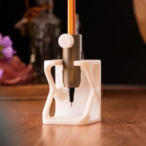 Könnte beinhalten: Ein weißer, 3D-gedruckter Stifthalter mit einem einzigartigen, offenen Design. Er hält einen grauen Stift mit einem orangefarbenen und gelben Bleistift. Die Spitze des Stifts ist sichtbar, und eine weiße Kappe ist angebracht. Der Halter steht auf einer Holzoberfläche.