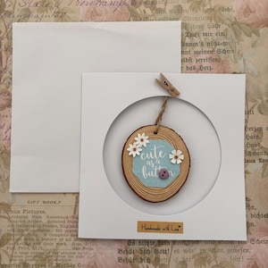 May include: A handmade wooden slice ornament with the text "cute as a button" on a light blue background, decorated with small white flowers and a purple button. The ornament is displayed in a white frame.