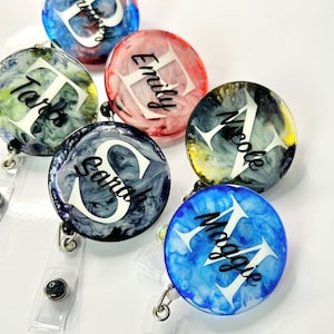 Könnte beinhalten: Fünf bunte, runde, personalisierte Ausweishalter mit einem weißen Initial und schwarzen Schriftzug. Die Namen sind Tara, Emily, Nicole, Sarah und Maggie.