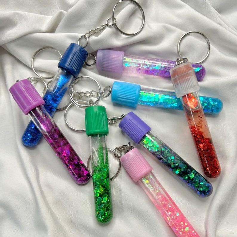 Pu&ograve; includere: Un set di sette tubi portachiavi pieni di glitter colorati con anelli portachiavi argentati. I tubi sono riempiti con glitter di diversi colori, tra cui blu, verde, viola, rosa e rosso.