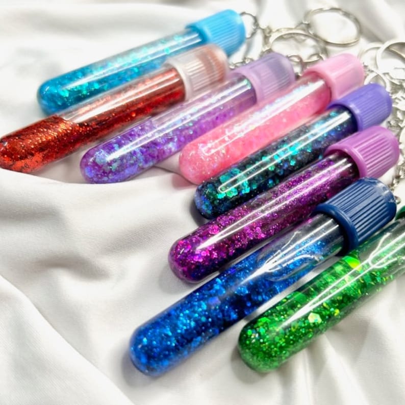 Pu&ograve; includere: Un set di otto tubi di vetro trasparente riempiti di glitter colorati, attaccati a portachiavi. I tubi sono tappati con vari colori, tra cui viola, rosa, blu e verde.
