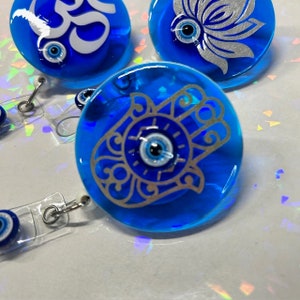 Puede incluir: Primer plano de varios portatarjetas de resina azul con varios diseños, incluyendo un símbolo Om, una flor de loto y una mano de Hamsa. Cada carrete tiene un cordón retráctil y un clip metálico.