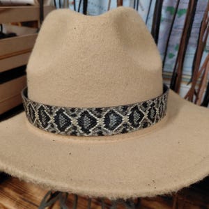 May include: A beige felt fedora hat featuring a wide brim and a pinched crown. The hat is accented with a decorative band around the base, displaying a repeating black, gray, and white diamond-shaped pattern that mimics a snakeskin texture.