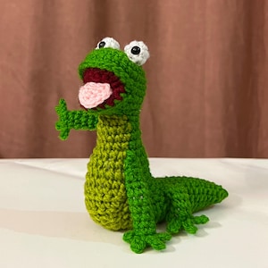May include: A green crocheted lizard figurine with a lighter green belly, a dark red open mouth, a light pink tongue, and two white eyes with black pupils. The lizard has four small feet and a long tail, standing in an upright position.