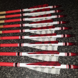 Pode incluir: Um conjunto de flechas de arco com hastes vermelhas e brancas. Cada flecha tem uma pena vermelha e o texto "Peter Fisher BCCB" impresso na haste. As flechas são numeradas de 1 a 7.