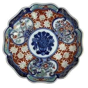 May include: A scalloped ceramic plate featuring a central blue and white floral motif surrounded by a rust-orange background with white flower patterns. The border includes dark blue sections with gold grid details and floral accents.