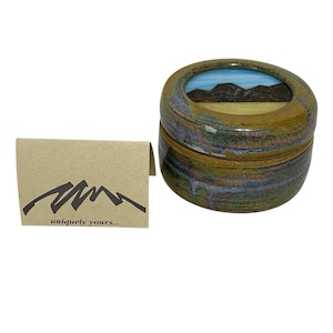 May include: A ceramic jar with a lid, featuring a landscape design. The jar is a mix of green, brown, and blue hues. A small card with a mountain graphic and the text "uniquely yours..." is next to the jar.