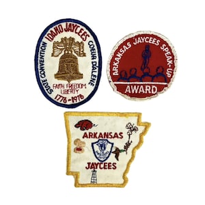 Puede incluir: Tres parches bordados con el diseño de la organización Jaycees. El primer parche es blanco con ribetes azules y rojos y presenta una campana con el texto "Idaho Jaycees State Convention Coeur d'Alene Faith Freedom Liberty 1776-1978". El segundo parche es rojo con ribetes blancos y presenta una persona hablando a un grupo de personas con el texto "Arkansas Jaycees Speak Up Award". El tercer parche es blanco con ribetes amarillos y presenta un mapa de Arkansas con el texto "Arkansas Jaycees" y varias imágenes, incluyendo un cerdo, una bola de algodón, un pato y una torre de perforación.