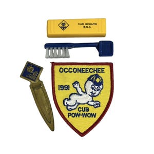 May include: A yellow and red embroidered patch with the text "Occoneechee 1991 Cub Pow-Wow". The patch features a cartoon baby wearing a blue hat. The patch is surrounded by a red border.