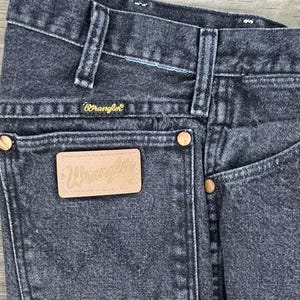 May include: Close-up of black denim Wrangler jeans. The jeans feature a classic five-pocket design, with copper-colored rivets and a leather patch on the back pocket. The Wrangler logo is visible on the patch and above the back pocket.