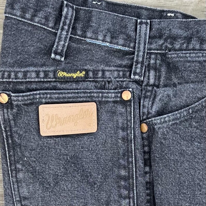 May include: Close-up of black denim Wrangler jeans. The jeans feature a classic five-pocket design, with copper-colored rivets and a leather patch on the back pocket. The Wrangler logo is visible on the patch and above the back pocket.
