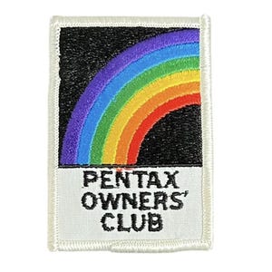 May include: Embroidered patch with a rainbow design on a black backdrop. The patch has a white border and the text "PENTAX OWNERS' CLUB" in black lettering.