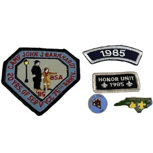 Può includere: Una collezione di toppe e spille vintage dei Boy Scout. Le toppe includono una toppa a forma di diamante con testo, una toppa curva con "1985" e una toppa rettangolare con "Honor Unit 1985". Le spille presentano un diamante e un contorno della Carolina del Nord.