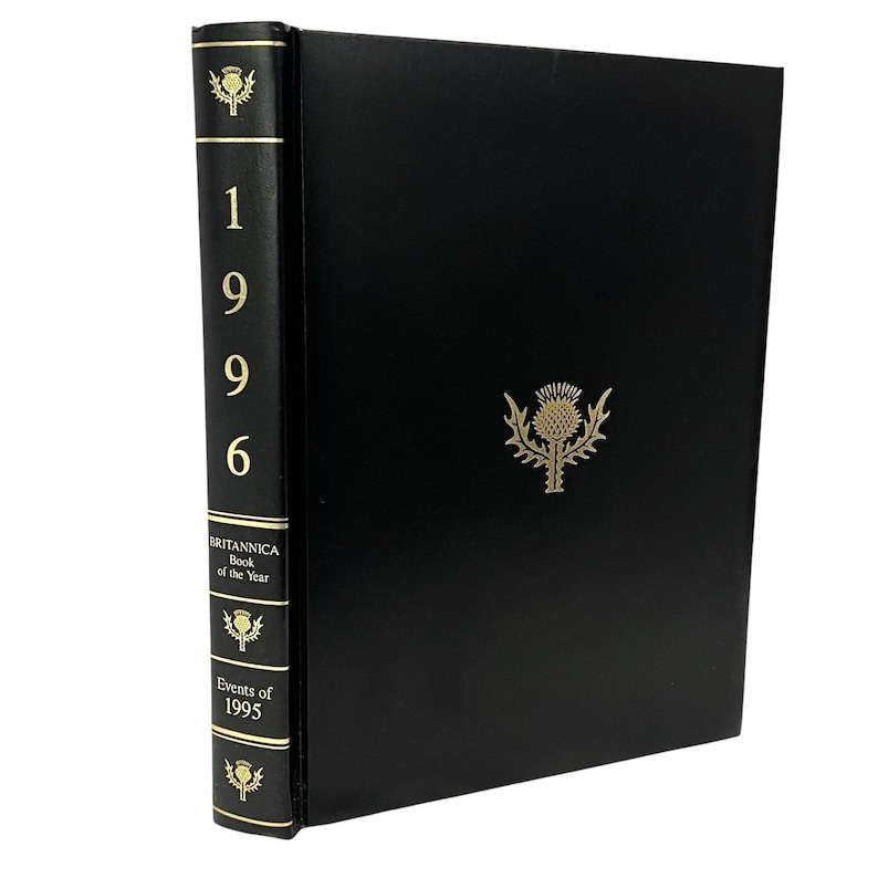 May include: A black hardback book, "Britannica Book of the Year Events of 1995". The spine displays the year "1996" in gold, with thistle emblems. The cover features a gold thistle emblem. A reference book.