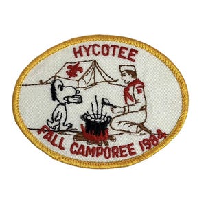 Może przedstawiać: Biała naszywka haftowana z żółtą obwódką. Naszywka przedstawia kreskówkowego psa, skauta gotującego nad ogniskiem i namiot. Tekst na naszywce brzmi "Hycotee Fall Camporee 1984".