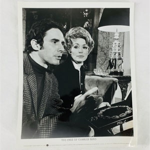 May include: Black and white promotional photo from "The Eyes of Charles Sand" featuring two people. A man in a tweed jacket points to a necklace in a box. The woman looks on. The film title is at the bottom.