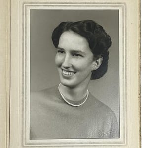 Puede incluir: Retrato en blanco y negro de una persona sonriente con un collar de perlas y un suéter. La persona tiene el pelo oscuro y mira hacia un lado. La fotografía está enmarcada en un borde claro.