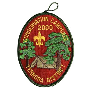 Puede incluir: Un parche bordado de forma ovalada roja con un borde verde. El parche presenta una tienda de campaña amarilla, dos árboles verdes, una flor de lis amarilla y el texto "Conservation Camporee 2000 Terrora District".