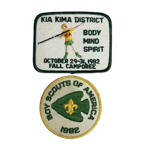 Może przedstawiać: Dwa haftowane naszywki, jedna z tekstem "KIA KIMA DISTRICT" i obrazkiem osoby rzucającej oszczepem, a druga z tekstem "BOY SCOUTS OF AMERICA" i rokiem 1982.