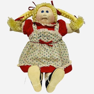 Könnte beinhalten: Eine Stoffpuppe mit blondem Wollhaar, das zu zwei Zöpfen geflochten und mit roten Bändern gebunden ist. Die Puppe hat blaue Augen, ein rundes Gesicht und trägt ein rotes Kleid mit einer cremefarbenen Blumenschürze, roter Schleife und Spitzenbesatz.