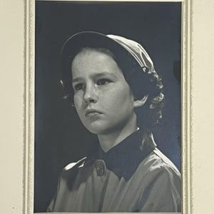 Puede incluir: Retrato en blanco y negro de una persona con sombrero y abrigo. La persona tiene pecas y mira hacia un lado. El sombrero tiene un ala oscura y una parte superior de color más claro. El abrigo tiene un cuello oscuro y un cierre de botón.