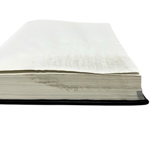 May include: A thick, rectangular book with a black cover and a stack of white pages. The pages are slightly aged, with some discoloration visible on the edges. The book is set against a white background.