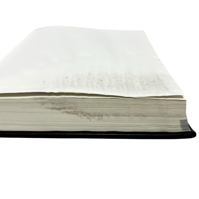 May include: A thick, rectangular book with a black cover and a stack of white pages. The pages are slightly aged, with some discoloration visible on the edges. The book is set against a white background.