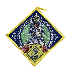 Peut inclure: Un écusson en forme de losange bleu et jaune avec une fusée et un logo de Cub Scout. Le texte sur l'écusson est "Galaxy of Scouting Cub Scout Pow Wow".