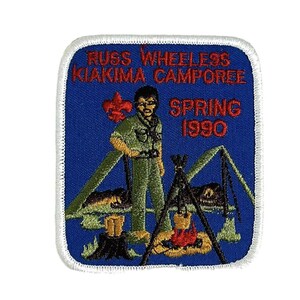 Könnte beinhalten: Ein blauer gestickter Aufnäher mit weißen Nähten. Der Aufnäher zeigt einen Cartoon-Pfadfinder, der vor einem Lagerfeuer und einem Zelt steht. Der Text auf dem Aufnäher lautet "Russ Wheeless Yakima Camporee Spring 1990".