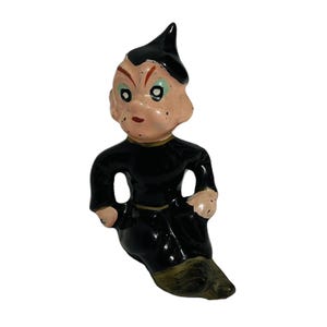 May include: A vintage ceramic figurine of a seated imp-like figure. The imp has a black pointed hat and black outfit with gold trim. The imp's face is peach-colored with red accents and green eyes. The figurine has a glossy finish.