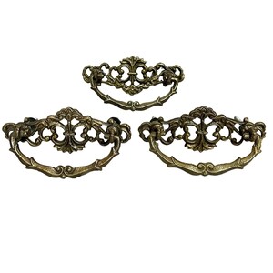 May include: Three matching drawer pulls made of antique brass-toned metal. Each pull features an ornate, symmetrical backplate with intricate scrollwork and a central fleur-de-lis motif, paired with a curved, swinging handle attached at two points.