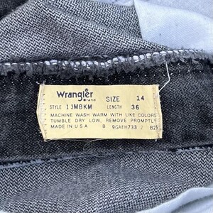May include: Close-up of a pair of dark denim Wrangler jeans. The tag reads "Wrangler" and includes the size 14 and length 36. The tag also includes washing instructions and "Made in USA".
