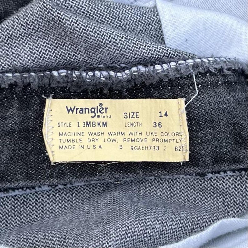 May include: Close-up of a pair of dark denim Wrangler jeans. The tag reads "Wrangler" and includes the size 14 and length 36. The tag also includes washing instructions and "Made in USA".