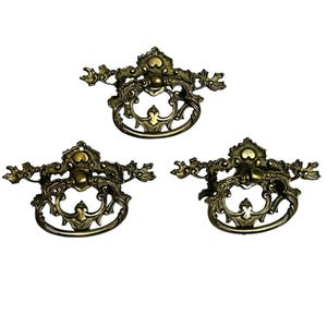 May include: Three matching antique brass drawer pulls featuring ornate, scrollwork backplates and swinging, circular drop handles. Each piece displays intricate metal detailing with a dark, aged patina finish and a central mounting point.