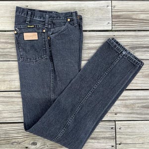 May include: Dark gray denim jeans with a classic five-pocket design. The jeans feature a leather patch with the "Wrangler" logo on the back pocket and brass rivets. The jeans are folded on a wooden surface.