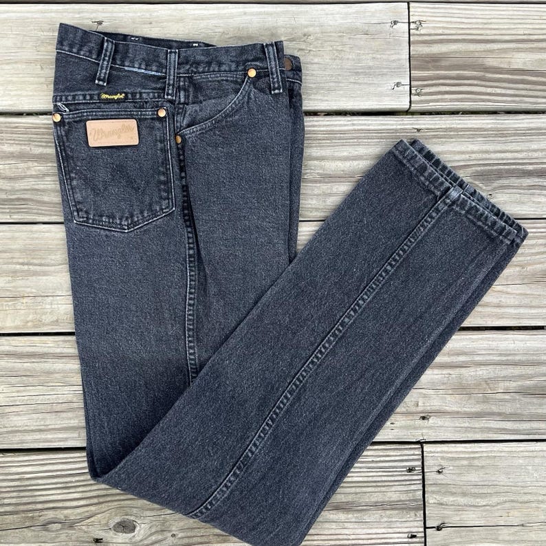 May include: Dark gray denim jeans with a classic five-pocket design. The jeans feature a leather patch with the "Wrangler" logo on the back pocket and brass rivets. The jeans are folded on a wooden surface.