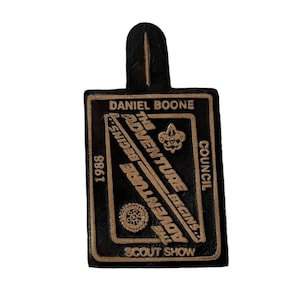 May include: A black leather patch with gold lettering that reads "Daniel Boone Council, The Adventure Begins, 1988, The Adventure Begins, Scout Show". The patch features a gold Boy Scout emblem.