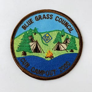 Puede incluir: Un parche bordado marrón con el texto "Blue Grass Council Cub Campout 2000". El parche presenta un cielo azul, hierba verde y dos tiendas de campaña con árboles a cada lado.